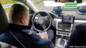 Zdjęcie kolorowe. Policjant ruchu drogowego siedzi wewnątrz radiowozu nieoznakowanego, wyposażonego w wideo rejestrator i zapisuje dane w notatniku służbowym.
