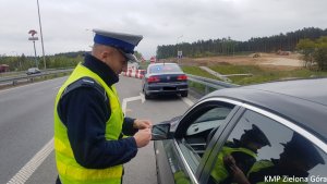 Zdjęcie kolorowe. Policjant ruchu drogowego stoi przy kontrolowanym samochodzie BMW, w ręce trzyma prawo jazdy kierującego. W tle widoczny policyjny nieoznakowany radiowóz.