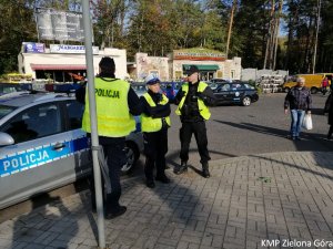 Policjanci stoją przy radiowozie i pilnują bezpieczeństwa na terenie parkingu cmentarza
