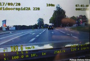Na zdjęciu widoczny jest ekran policyjnego videoradaru z zapisem prędkości samochodu 103 km/h, w tle droga z samochodem