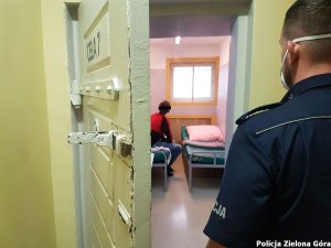 Mężczyzna siedzący w celi policyjnego aresztu. W otwartych drzwiach celi stoi policjant w mundurze.